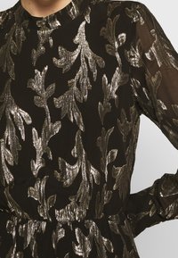 Robe en tissu noir ornée de motifs de feuilles argentées métalliques, manches longues et col haut. Matière texturée et légère avec une taille cintrée.