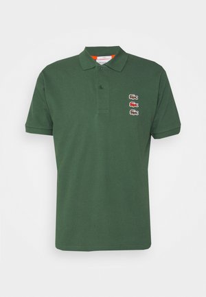 Ciemnozielona koszulka polo z krótkim rękawem, plisą na dwa guziki i trzema wyszywanymi logotypami krokodyla w kolorach brązowym, czerwonym i brązowym na lewej stronie klatki piersiowej.
