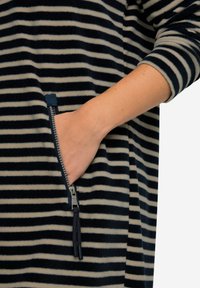 Raidallinen fleece-poolo, jossa on navy- ja kreemivalkoiset vaakasuorat raidat, sivuvetoketjullinen tasku, jossa on tupsu, sekä ranteen pituinen hiha.