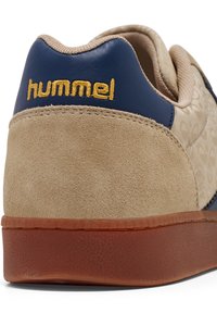 Beige suede sneaker with blue accents, textured rubber sole, and gold "hummel" logo at the heel. Features a subtle pattern on the upper.