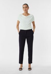 Světle zelený draped top s krátkými rukávy, doplněný o přiléhavé tmavě modré kalhoty. Černé sandály s otevřenou špičkou dotvářejí vzhled.