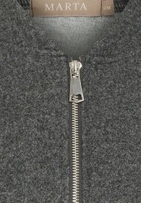 Grå uldblandet jakke med lynlåslukning foran. Sølvfarvet lynlåsdetalje går fra kraven til kanten og suppleres af en tekstureret finish.