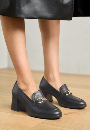 Mocassins à talons en cuir noir avec un bout arrondi, un détail doré et un talon bloc, posés sur un sol en bois.