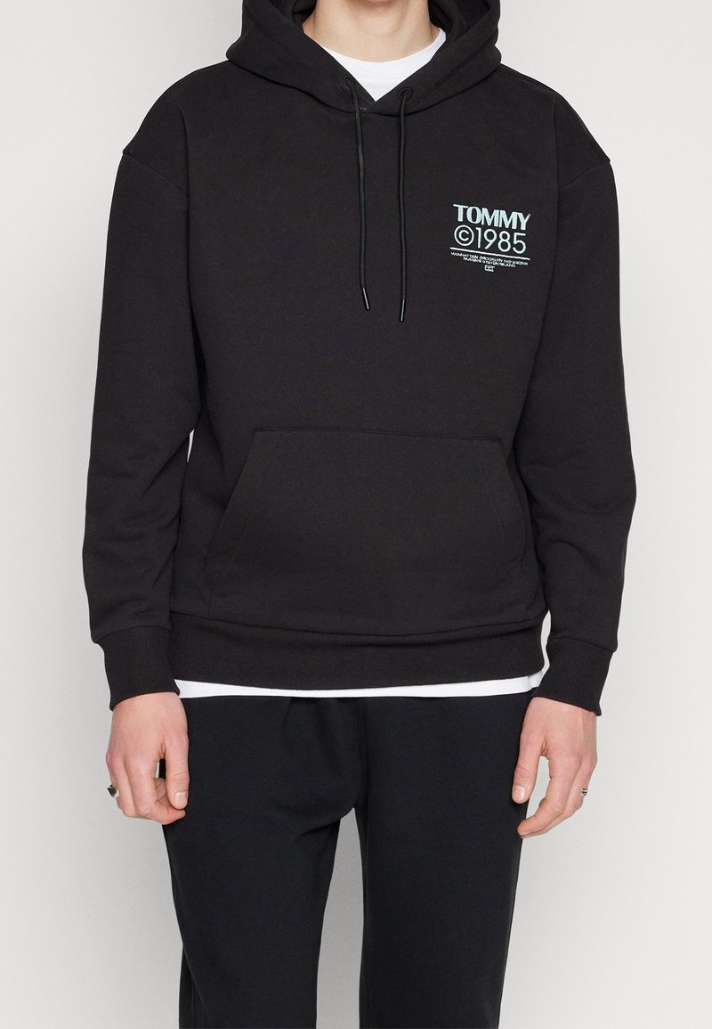 Sweat à capuche noir en mélange de coton, avec une poche avant, des poignets côtelés et une capuche avec cordon de serrage. Logo "TOMMY 1985" en couleurs contrastantes.