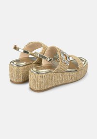 Sandali con zeppa beige, con parte superiore in tessuto intrecciato, accenti metallici e una fibbia decorata di gioielli. La suola piattaformata è texturizzata e rialzata.