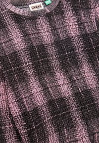 Gros plan d’un tissu texturé à motifs rose et noir avec une étiquette GUESS Originals et une étiquette de la collection éco près de l’encolure.