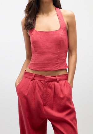 Woman wearing a sleeveless, square-neck red crop top paired with high-waisted, pleated red pants with hands in pockets.
