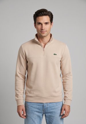 Uomo che indossa una felpa beige con zip a quarto e logo di coccodrillo verde, e jeans azzurri, in piedi su uno sfondo grigio uniforme.