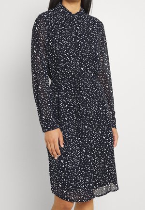 Femme portant une robe bleu marine à manches longues avec un motif de pois blancs dispersés et une ceinture nouée à la taille, debout devant un fond uni.