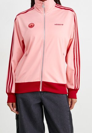 Rosa und rote Adidas-Trainingsjacke mit Reißverschluss und Streifen an den Ärmeln, getragen mit dunklen grauen Hosen, Hände entspannt an den Seiten.