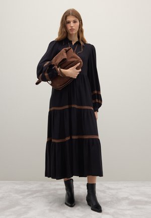 Vestido negro de manga larga con diseño de volantes y detalles de costuras marrones contrastantes, combinado con un bolso de cuero marrón y botines negros.