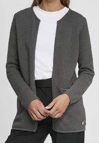 Gray textured cardigan with a straight cut, round neckline, no buttons, two front pockets, and a small logo accent. Worn over a white shirt.