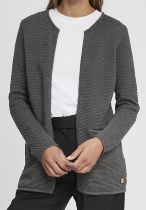 Grauer, strukturierter Cardigan mit geradem Schnitt, rundem Ausschnitt, ohne Knöpfe, zwei Fronttaschen und einem kleinen Logo-Akzent. Darunter wird ein weißes Shirt getragen.
