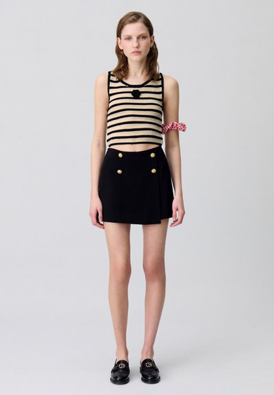 Top corto a rayas blancas y negras, falda mini negra con botones dorados y mocasines negros. Acento a cuadros rojos y blancos en el brazo.
