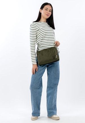 Grüne rechteckige Umhängetasche mit glatter Textur und Logo. Das Model trägt ein gestreiftes Langarmshirt und blaue Jeans mit weitem Bein.