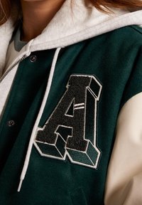 Vihreä ja kerma varsity-takki, jossa valkoinen huppari. Takissa on etuosassa näkyvä kohokuvioitu musta kirjain "A" ja nepparikiinnitys.