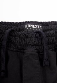 Mustahdud musta, jossa on nyöriysitys, jossa on "HONESTY RULES" -tiketti. Materiaali näyttää sileältä ja siinä on hieman kiiltoa.