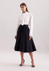 Camicia bianca a maniche lunghe con collo annodato, abbinata a una gonna nera plissettata a lunghezza ginocchio e sandali neri con tacco alto e cinturini.