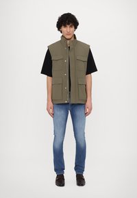 Khaki mouwloos vest met rits aan de voorkant, twee borstzakken en twee zijzakken, gedragen over een zwart T-shirt en blauwe jeans.