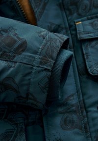 Teal kviltad jacka med svarta maskintrycksmönster. Har ribbade cuffs och en ficka, tillverkad av glatt, hållbart tyg.