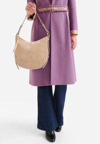 Borsa hobo in suede beige dalla forma curva, dotata di tracolla in pelle color cuoio e dettagli in metallo tono oro discreto. Indossata con un cappotto viola e jeans scuri.