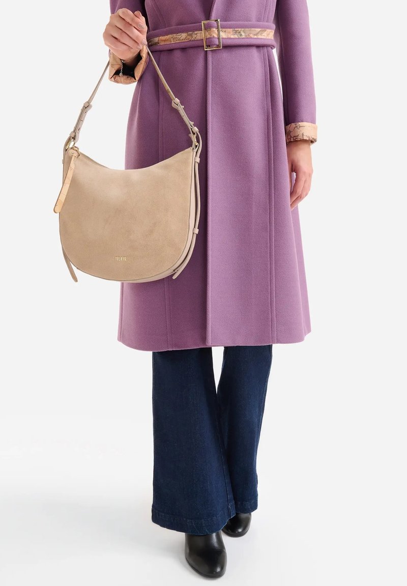 Borsa hobo in suede beige dalla forma curva, dotata di tracolla in pelle color cuoio e dettagli in metallo tono oro discreto. Indossata con un cappotto viola e jeans scuri.