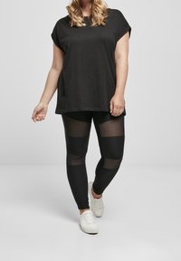 Czarna, oversize'owa koszulka z podwiniętymi rękawami, zestawiona z czarnymi legginsami z przezroczystymi panelami i prążkową teksturą, noszona z białymi trampkami.