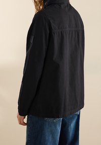 Black jacket made of lightweight fabric, featuring a high collar, visible seams, and a rectangular patch on the left sleeve. Worn over blue jeans.