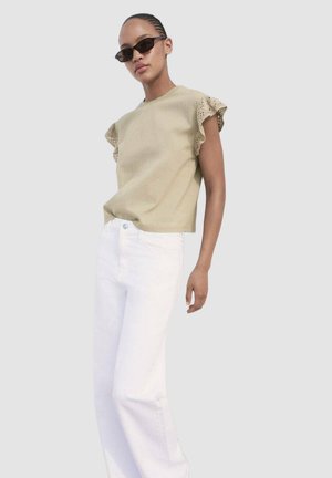 Licht olijfgroene blouse met gerimpelde kantmouwen, gecombineerd met witte denim broek. De outfit heeft een relaxte pasvorm en een soepele textuur.