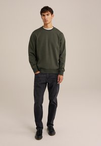 Sudadera verde oscura con cuello redondo, mangas largas y puños de canalé, combinada con jeans oscuros y zapatos negros.