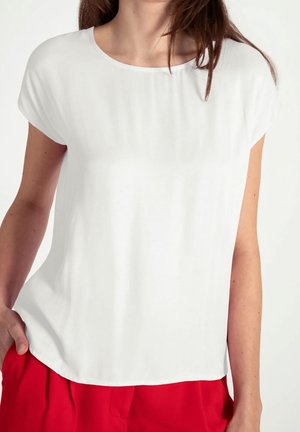 Mujer con una camiseta blanca lisa de manga corta y pantalones rojos brillantes, con la mano en el bolsillo sobre un fondo claro.