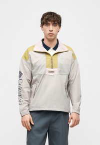 Νεαρός άνδρας που φοράει ένα pastel αμυντικό μπουφάν Columbia με φερμουάρ μπροστά και λογότυπο στο αριστερό μανίκι, στέκεται μπροστά σε έναν απλό λευκό φόντο.