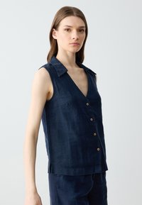 Marineblauwe mouwloze linnen blouse met een v-hals, kraag en houten knopen. Heeft een subtiele textuur en zijslits voor comfort.