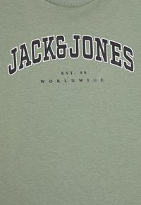 Gaiši zaļš kokvilnas T-krekls ar melniem burtu uzrakstiem "JACK & JONES", kam zemāk mazākā drukā ir teksts "EST. 90" un "WORLDWIDE".