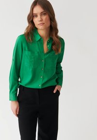 Cămașă verde cu nasturi, confecționată dintr-un material neted, având două buzunare la piept și detalii cu nasturi, asortată cu pantaloni negri.