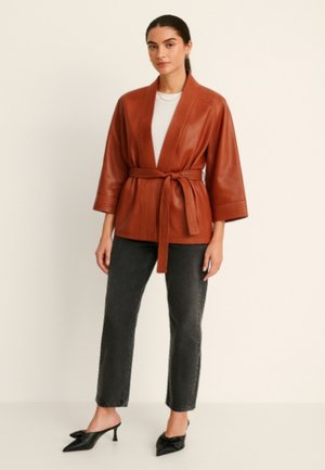 Veste en cuir marron de style kimono avec une ceinture à nouer, détails côtelés sur les manches, associée à un jean sombre et des mules à talons noires.