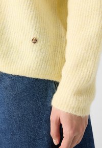Gelber, weicher Strickpullover mit gerippten Bündchen und einem kleinen goldenen Akzent am Saum. Wird über Denim mit strukturiertem Finish getragen.