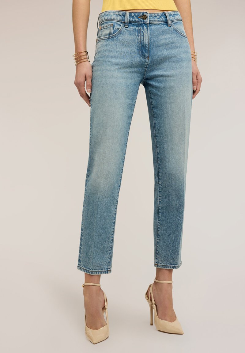 Frau trägt hellblaue Jeans mit geradem Bein, gelbes ärmelloses Oberteil, goldene Armbänder und beige High-Heel-Sandaletten mit Knöchelriemen.