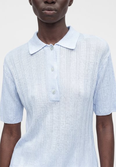 Femme portant un polo en maille bleu clair à manches courtes avec une texture côtelée verticale et trois boutons blancs sur la patte de boutonnage.