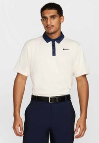 Koszulka polo z krótkim rękawem w kremowym kolorze z granatowym kołnierzykiem i czarnym logo Nike po lewej stronie piersi, zestawiona z granatowymi spodniami i czarnym paskiem.