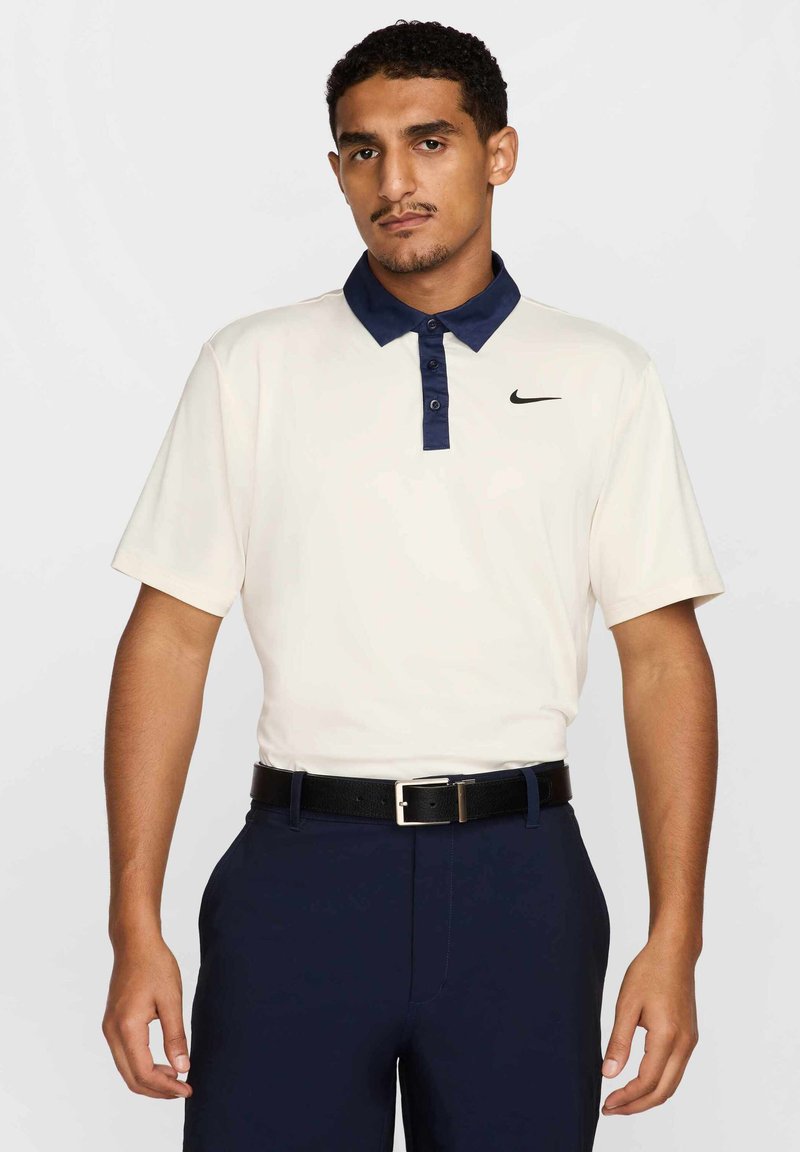 Koszulka polo z krótkim rękawem w kremowym kolorze z granatowym kołnierzykiem i czarnym logo Nike po lewej stronie piersi, zestawiona z granatowymi spodniami i czarnym paskiem.