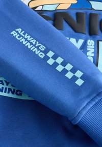 Modrý rukáv mikiny se světle modrým textem "ALWAYS RUNNING" a kostkovaným vzorem nad částečným kresleným motivem.