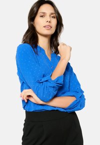 Camicia button-up blu royal con un motivo testurizzato, maniche arrotolate e colletto arrotondato, abbinata a pantaloni neri.