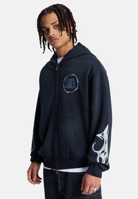 Νεαρός άνδρας που φοράει σκούρο φούτερ με φερμουάρ, με κυκλικό λογότυπο στο στήθος και σχέδιο λευκής φλόγας στο αριστερό μανίκι, στέκεται μπροστά σε λευκό φόντο.