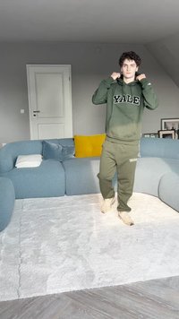 Fleece verde com o estampado "YALE", calças de fato combinando, sapatos creme. Quarto com sofá azul e almofada amarela sobre um tapete branco e texturizado.