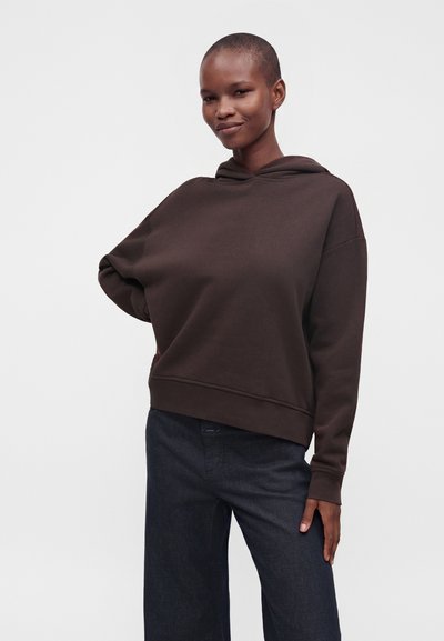 Personne aux cheveux courts portant un sweat à capuche marron foncé et un pantalon large foncé, debout et souriant devant un fond uni clair.