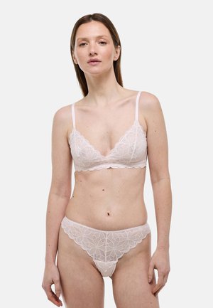 Femme portant un ensemble assorti de soutien-gorge en dentelle rose clair et de culotte, debout devant un fond gris uni, face à l'avant avec une expression neutre.
