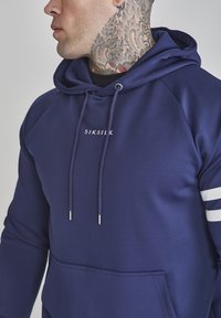 Tengerészkék kapucnis pulóver, kenguruzsebbel, fehér csíkokkal az ujjakon és ezüst végű húzózsinórral; az SIKSILK márka logója a mellrészen található.