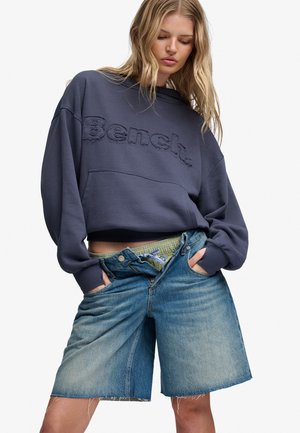 Ung person iført mørk oversized sweatshirt med "Bench" logo og løstsiddende, falmet denimshorts med frynset kant og synligt undertøjsbælte.