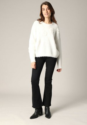 Un pull blanc duveteux avec des détails texturés floraux, associé à un jean noir évasé et des bottines noires. Fond neutre.